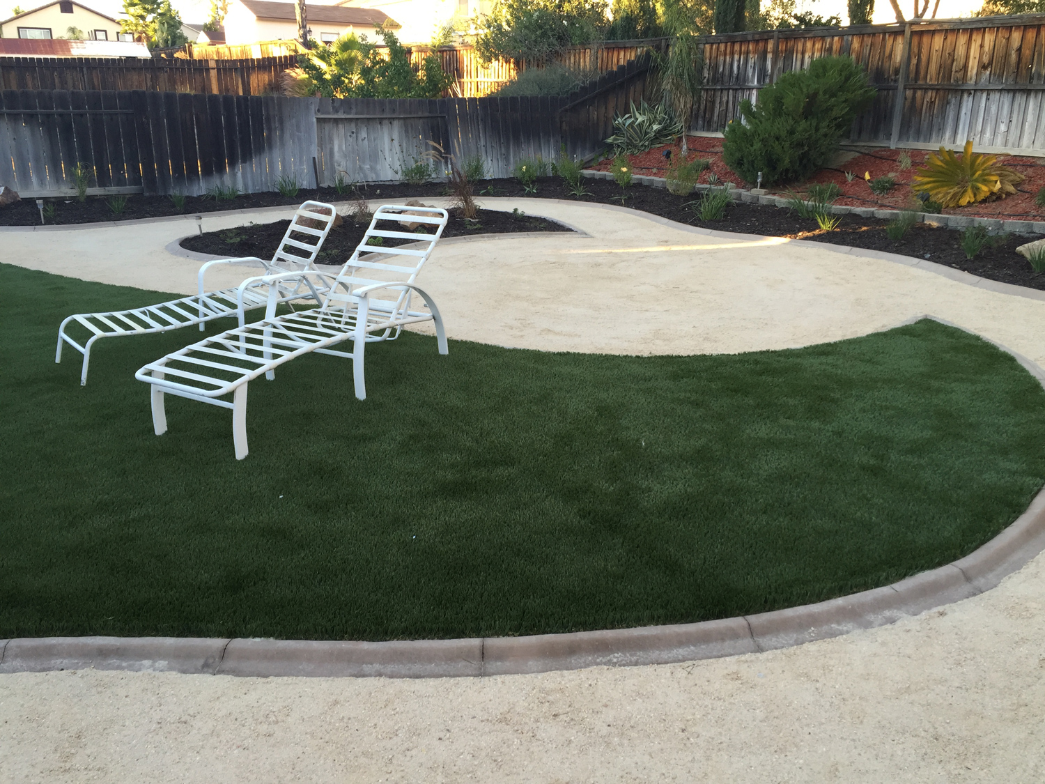 Backyard landscaping with white lawn chairs, lush dark green grass and mulch beds with plants.